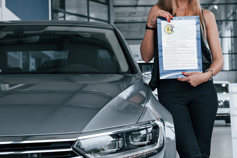 Coche gris con mujer al lado con la garantía del vehículo entre las manos. 