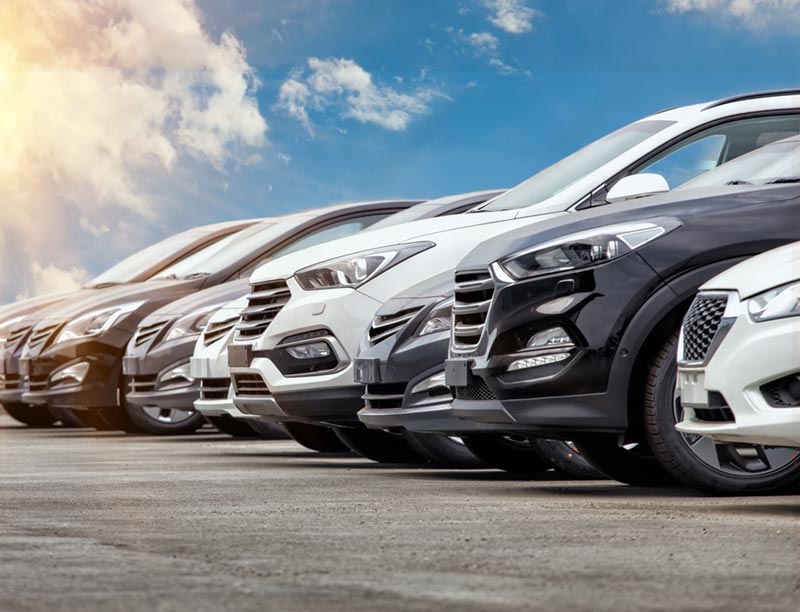 Parte delantera de fila de coches con un cielo azul con nubes y rayos de sol.